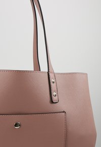 Sac à main texturé rose pâle avec des fines bretelles, des rivets argentés sécurisant la sangle, et une poche avant avec une fermeture à bouton argenté.