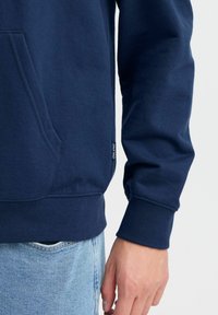 Marineblauwe hoodie met een kangoeroezak, ribgebreide manchetten en gestructureerde stof. Beschikt over een klein merkje aan de zijkant.