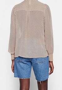 Blusa beige trasparente con pois marroni, maniche lunghe e dettaglio del yoke sul retro, abbinata a shorts in denim blu chiaro con orli sfrangiati.