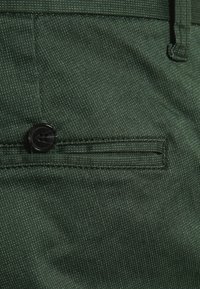 Gröna tygbyxor med en strukturerad väv, en ficka med lock som stängs med en svart knapp, och en matchande bältesöppning.