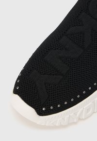 Pantof sport negru tricotat cu partea superioară texturată, prevăzut cu ținte argintii de-a lungul cusăturii și o talpă de cauciuc albă cu brand.