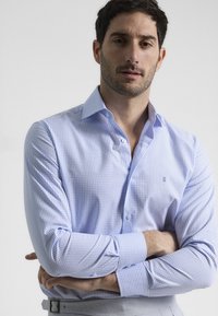 Camisa de vestir a rayas en azul claro con cuello abotonado, que presenta sutiles patrones de cuadrícula en blanco y morado y mangas largas.