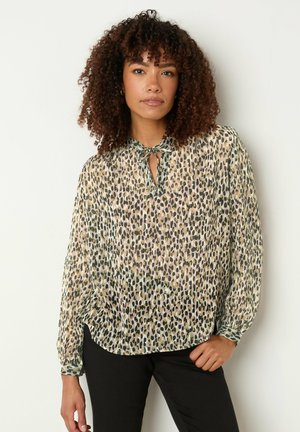 Kvinde med krøllet hår iført en langærmet, mønstret bluse med bindebånd ved halsen og sorte bukser, stående foran en ensfarvet lys baggrund.