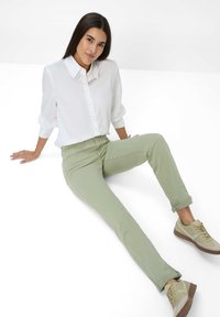 Weißes Hemd mit Kragen, hochgekrempelte Ärmel; hellgrüne Slim-Fit-Hose; hellbraune Sneaker mit Schnürsenkeln und braunen Akzenten; sitzende Pose, neutraler Hintergrund.