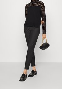 Haut noir à manches longues avec motif en résille, associé à un pantalon noir slim et des mocassins noirs brillants. Accessoire : un petit sac à main noir à chaîne dorée.