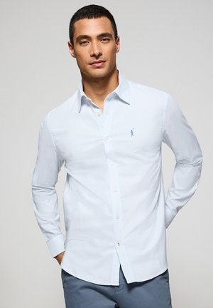 Camisa de botones a rayas azul claro con cuello clásico, mangas largas y un pequeño logo bordado en el lado izquierdo. Textura de tela suave.