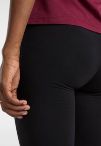 Närbild av en person som bär svarta leggings och en vinröd skjorta, med vänsterhanden vilande nära låret.