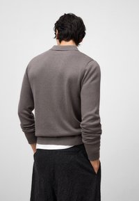Maglione grigio a maglia con colletto e polsini a coste, indossato sopra una maglietta bianca, abbinato a pantaloni neri texturizzati. Vista posteriore.