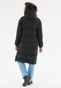 Schwarze Pufferjacke mit Kapuze, lange Ausführung, mit vertikalen Steppabschnitten. Kombiniert mit hellblauen Jeans und schwarzen Stiefeln.