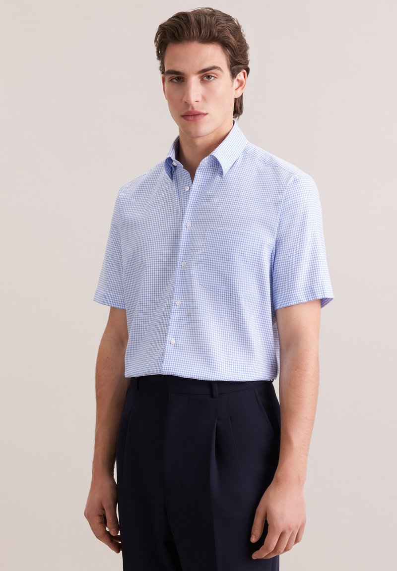 Jeune homme portant une chemise à manches courtes bleu clair à carreaux et un pantalon sombre, debout devant un fond beige uni.