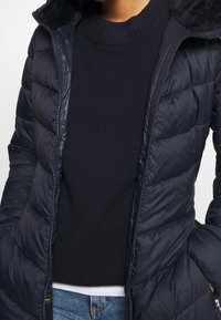 Marineblaue Steppjacke mit gestepptem Design und Reißverschluss, ausgestattet mit einem weichen, pelzgefütterten Kragen, getragen über einem dunklen Pullover und blauen Jeans.