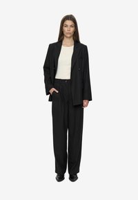 Kvinde iført en sort oversized blazer, brede bukser, en hvid ribbet top og sorte spidse sko, stående med den ene hånd i lommen.