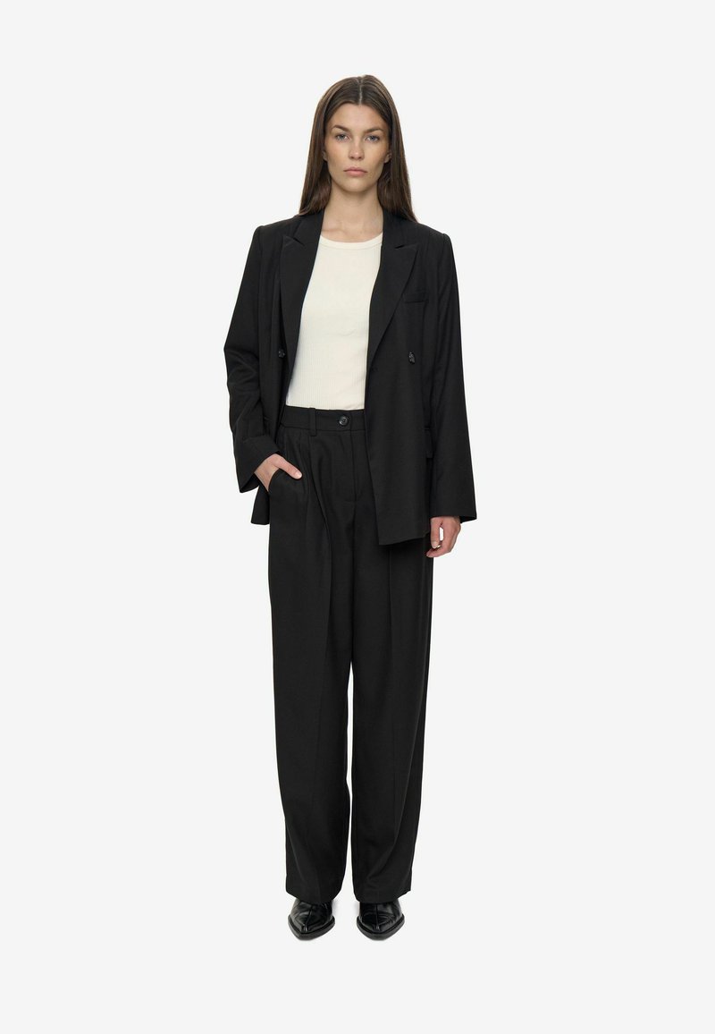 Kvinde iført en sort oversized blazer, brede bukser, en hvid ribbet top og sorte spidse sko, stående med den ene hånd i lommen.