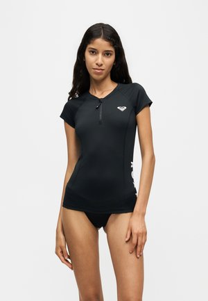 Giovane donna che indossa una maglia da bagno nera a maniche corte con zip e piccolo logo sul petto e slip bikini neri, in piedi davanti a uno sfondo semplice.