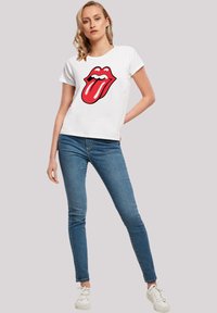White cotton t-shirt with a red and black graphic design of lips and a tongue, paired with blue jeans and white sneakers.