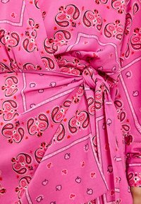 Le tissu rose présente un motif paisley avec des accents rouges et de petits cœurs. Le matériau est lisse, et le design inclut un détail de taille nouée.