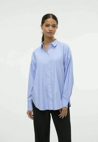 Camicia azzurra chiara a maniche lunghe con strisce bianche verticali, colletto e maniche con polsini. Indossata con pantaloni neri.