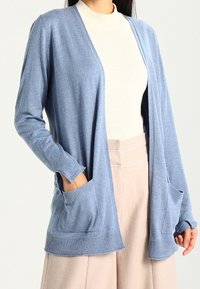 Ljusblå cardigan i mjukt, stickat tyg med v-ringning. Har två påsydda fickor och ribbade muddar, kombinerad med en cream-färgad topp.