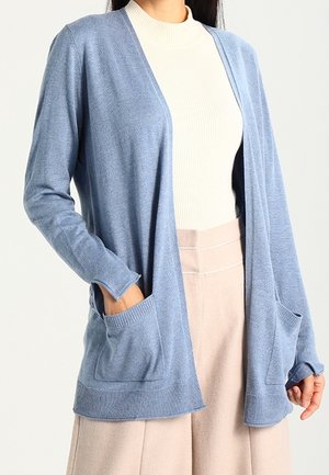 Donna con un cardigan aperto di colore azzurro chiaro sopra un maglione bianco a collo alto a coste e pantaloni beige chiaro, con le mani nelle tasche del cardigan.