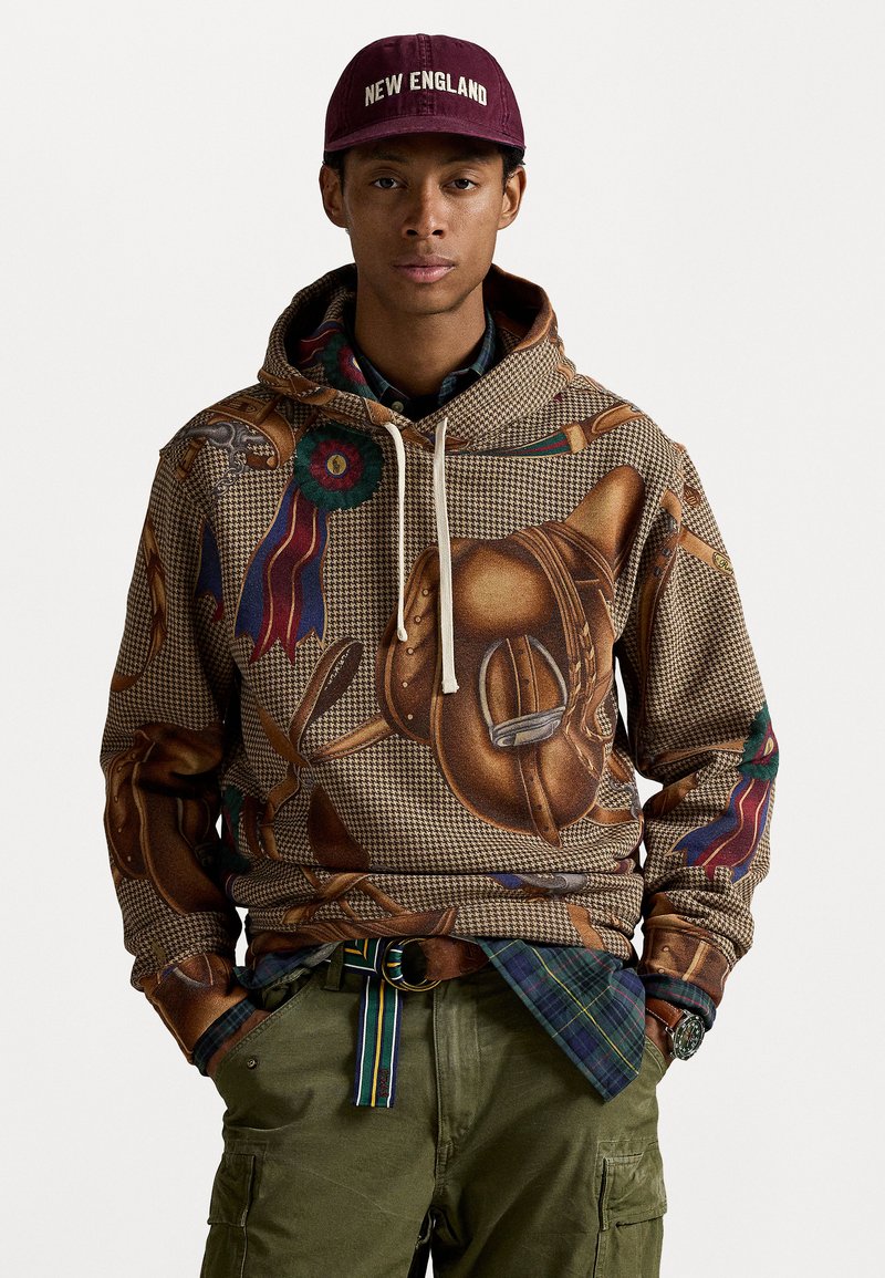 Homem com um boné bordô "New England", um hoodie castanho padrão com equipamento equestre, calças de carga verdes, camisa de xadrez e um cinto riscado, com as mãos nos bolsos.