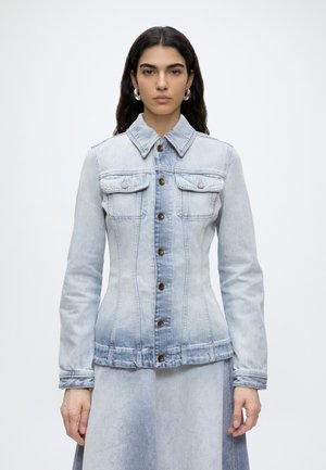 Femme aux longs cheveux foncés porte une veste en jean bleu clair cintrée avec des boutons en laiton et une jupe assortie sur un fond uni.