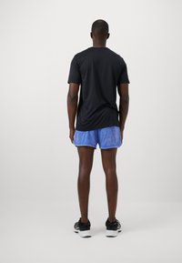 Schwarzes kurzärmliges Hemd, blaue gemusterte Shorts und schwarze Turnschuhe. Schlichtes Design, leichtes Material und taillierte Passform. Von hinten betrachtet.