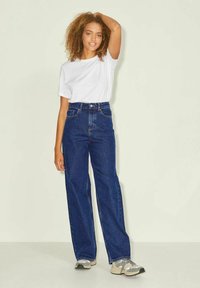 Mlada žena s kodrastimi lasmi pozira v beli majici, širokih modrih jeansih visoke pasu in sivih supergah na enobarvni podlagi.