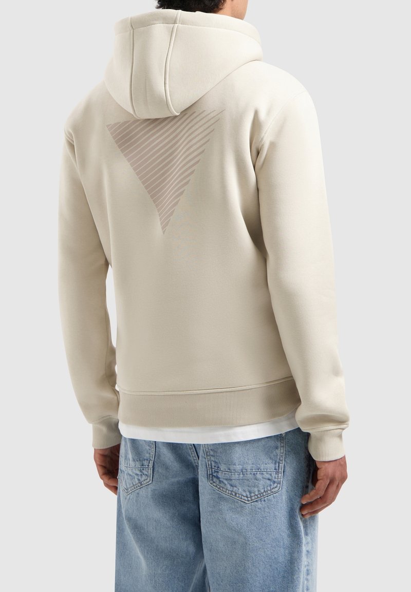 Beige hoodie met een gestructureerd driehoekig ontwerp op de achterkant. Voorzien van een capuchon en geribbelde manchetten. Draagt met lichtblauwe jeans.