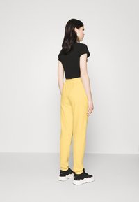 Schwarzes, verkürztes T-Shirt und gelbe Sweatpants. Die Hose hat einen elastischen Bund und schmal zulaufende Beine; kombiniert mit schwarzen Turnschuhen mit weißen Sohlen.