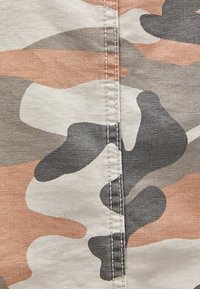 Tessuto mimetico con forme irregolari in beige, marrone chiaro, grigio e nero. Cuciture visibili lungo il centro con una superficie testurizzata.