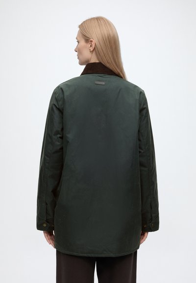 Femme aux longs cheveux blonds portant une veste vert foncé avec un col marron, vue de dos sur un fond clair uni.