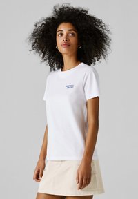 Žena s objemnými kučeravými vlasmi, oblečená v bielom tričku "Red Bull Racing" a béžovej sukni, stojaca na jednoduchom sivom pozadí.