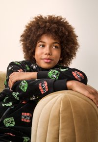 Black fleece onesie with colorful gaming-themed patterns, featuring a zip-up design and ribbed cuffs, worn while resting on a beige couch.