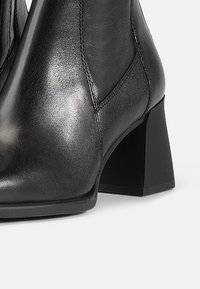 Bottines en cuir noir avec une finition brillante, comportant un talon carré et des détails de couture visibles. Texture lisse et ajustement confortable.
