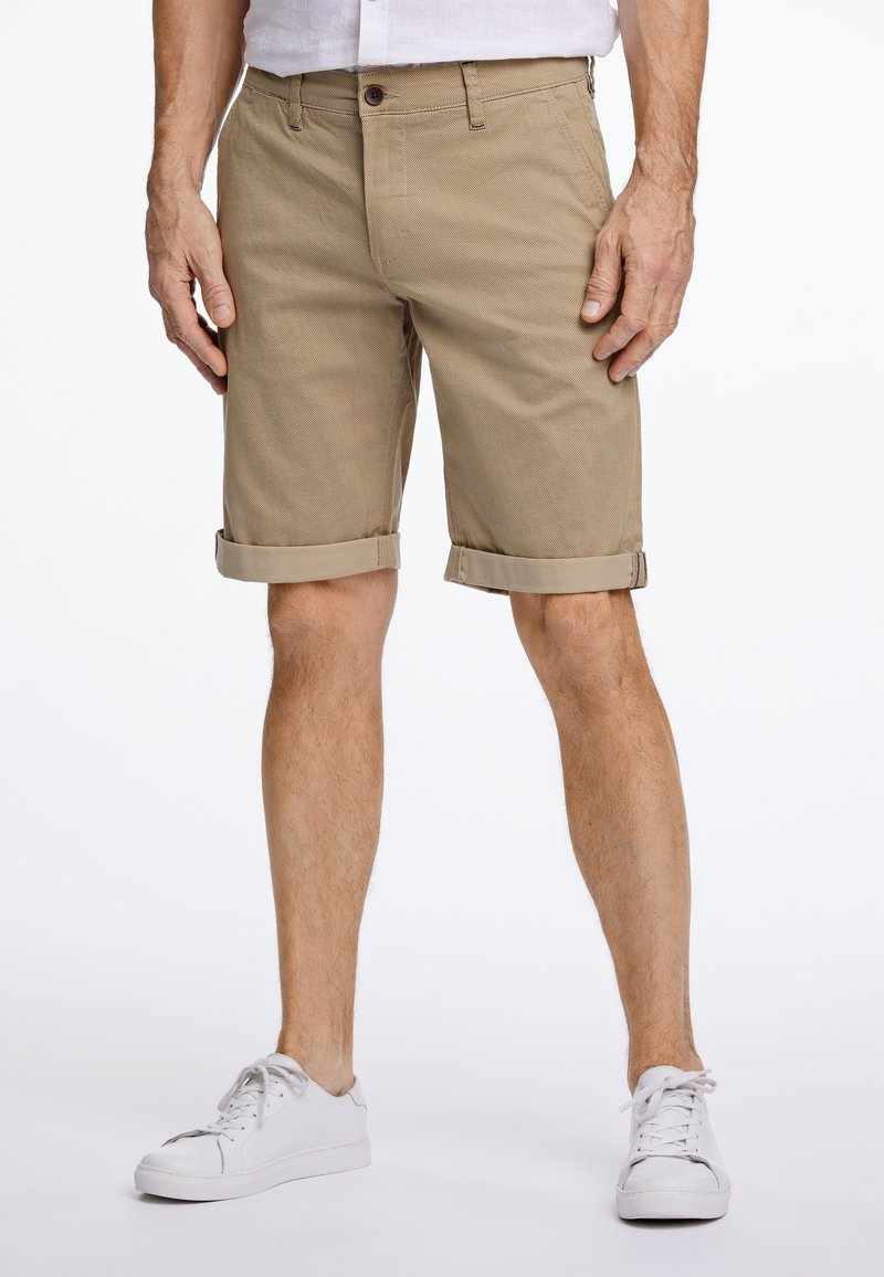 Mann trägt beige hochgekrempelte Shorts und weiße Turnschuhe, steht vor einem schlichten weißen Hintergrund.