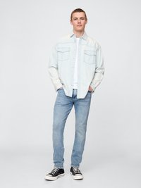 Camisa de mezclilla azul clara con dos bolsillos frontales, encima de una camiseta blanca. Llevada con jeans azul claros y zapatillas negras.