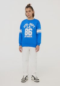 Felpa blu con stampa e righe bianche, abbinata a jeans bianchi strappati. Le sneaker in bianco e nero completano l'outfit.