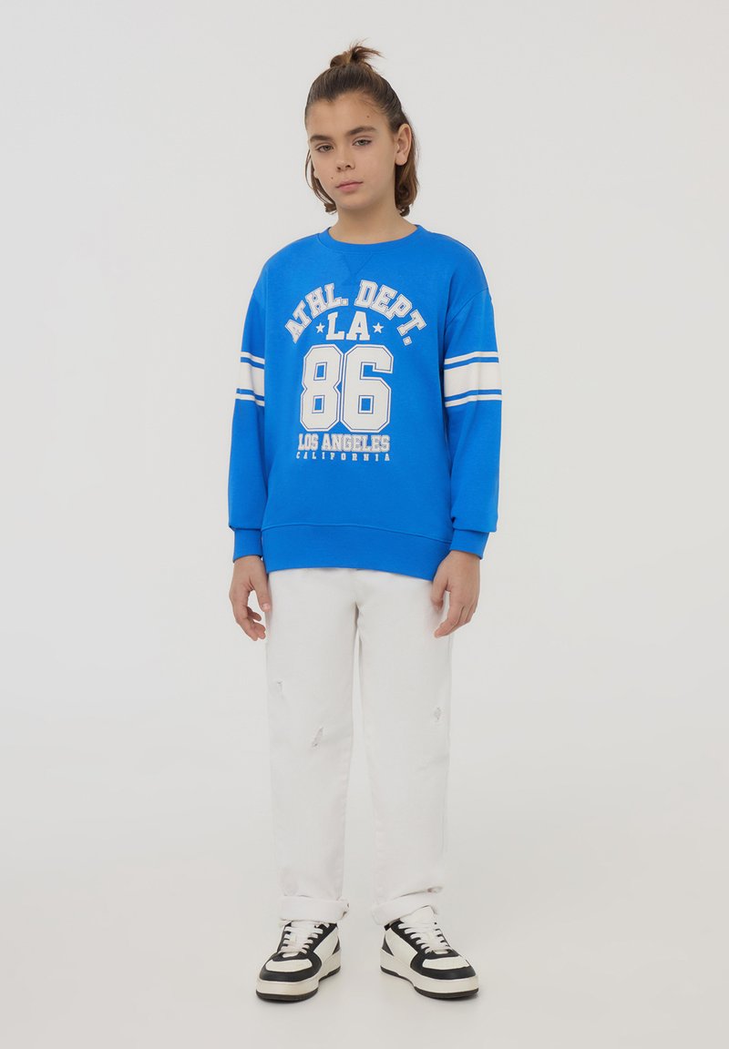 Felpa blu con stampa e righe bianche, abbinata a jeans bianchi strappati. Le sneaker in bianco e nero completano l'outfit.