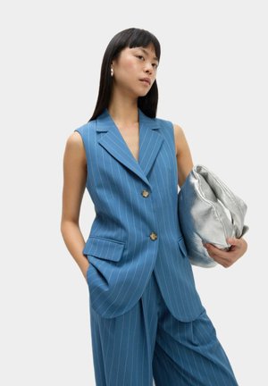 Chaleco sin mangas a rayas azules con dos botones y bolsillos laterales, combinado con pantalones a juego. Sosteniendo un bolso metálico plateado.