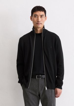 Black zip-up jacket with a high collar, made of a soft material. Features a silver zipper and subtle logo on the chest. Paired with grey trousers.