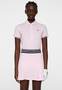 Rosa pikétröja med korta ärmar, med en svart logotyp på bröstet. Tillsammans med en veckad rosa kjol och svart-vita randig midja.