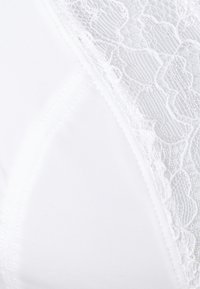 Bílá podprsenka z hladkého materiálu s krajkovým překryvem nahoře, zdůrazňující květinové vzory. Design zahrnuje jemné švy.