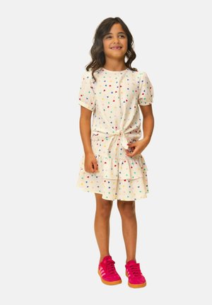 Young girl smiling, wearing a white dress with colorful heart patterns and bright pink sneakers, standing against a plain background.