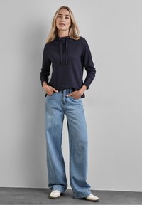 Granatowa bluzka z długim rękawem, z kapturem i sznurkiem, w połączeniu z jasnoniebieskimi szerokimi spodniami dżinsowymi i białymi trampkami. Swobodny, wygodny fason.