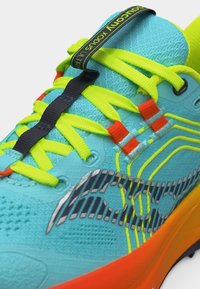 Κλείσιμο εικόνας μπλε αθλητικού παπουτσιού με κίτρινες κορδόνια, πορτοκαλί λεπτομέρειες, μαύρο λογότυπο και ιμάντα με κείμενο που γράφει "Saucony Xodus Ultra".