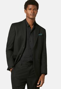 Veste de costume noire sur mesure à deux boutons, portée avec une chemise de costume noire assortie. Un mouchoir de poche teal est visible dans la poche de la veste.