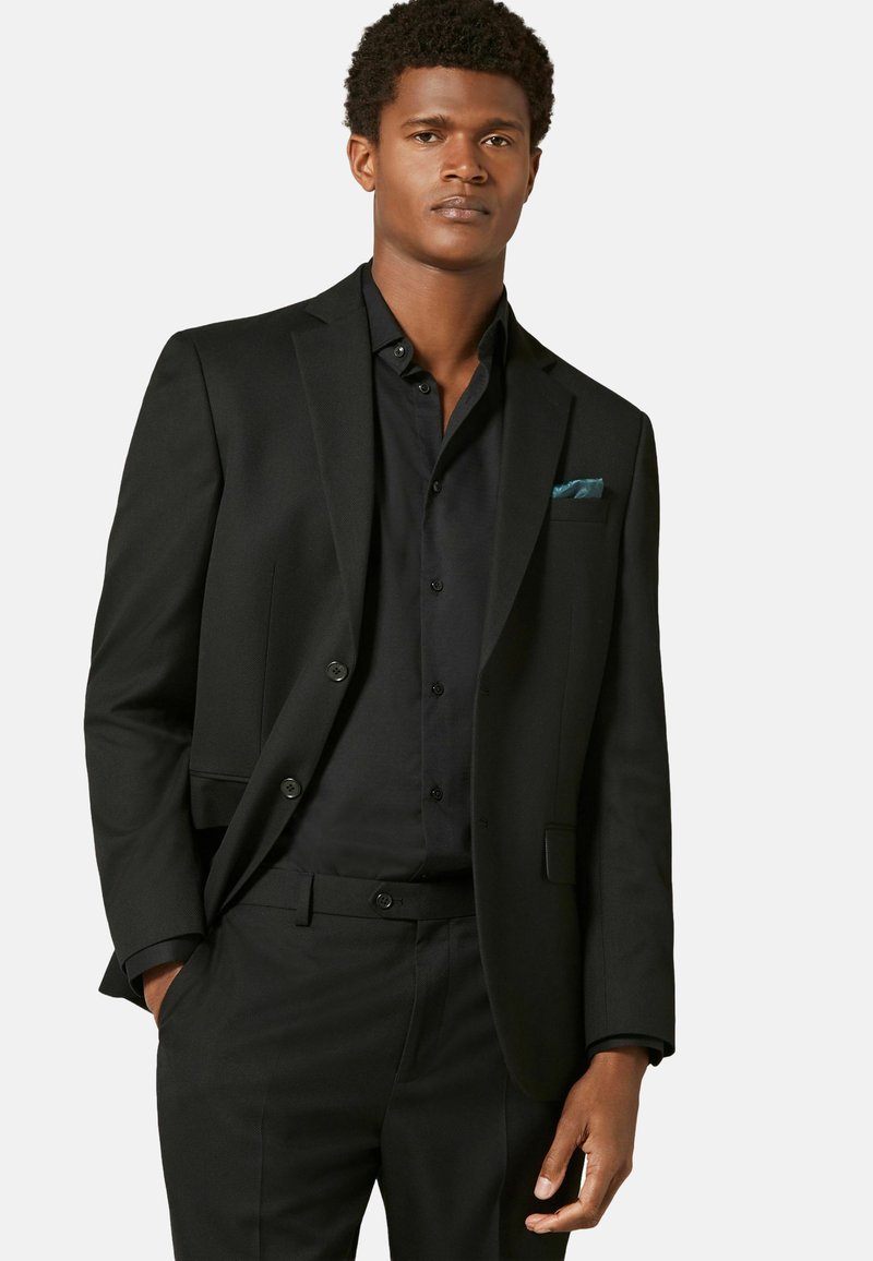 Veste de costume noire sur mesure à deux boutons, portée avec une chemise de costume noire assortie. Un mouchoir de poche teal est visible dans la poche de la veste.
