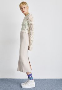 Witte gebreide crop sweater met groene accenten, beige aansluitende rok met een split aan de zijkant, gelaagd met paarse bloemenpatroon sokken en hoge witte sneakers.
