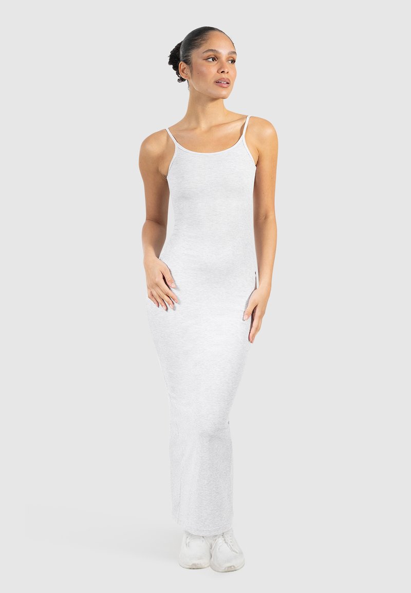 Robe maxi blanche en coton avec des bretelles fines, coupe ajustée et texture côtelée. Modèle debout en baskets blanches devant un fond gris clair.