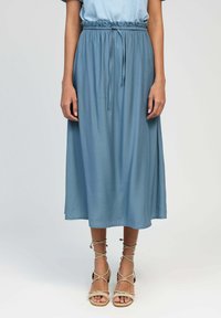 Jupe bleu clair, arrivant aux genoux, avec une taille élastique et un détail à nouer. Fabriquée en tissu léger, elle présente une silhouette fluide et un léger éclat.